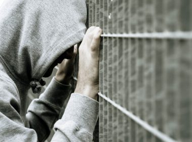 Male wearing hooded sweatshirt leaning against a prison fence