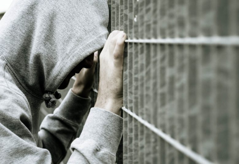Male wearing hooded sweatshirt leaning against a prison fence