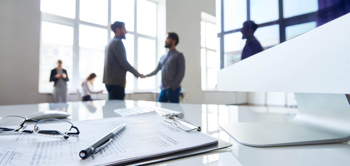 A signed contract sits on a desk and two men shakes hands in the background.