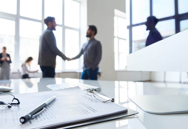 A signed contract sits on a desk and two men shakes hands in the background.