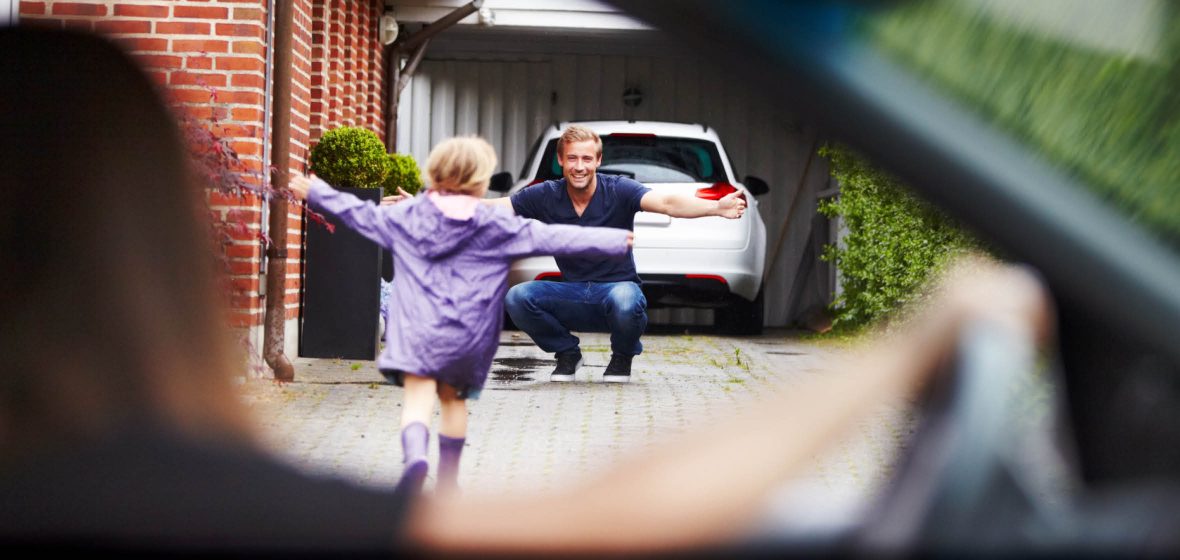 Child runs up a driveway towards a man while a woman sitting in a car watches them.