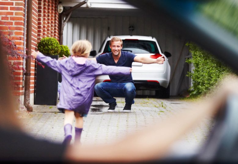 Child runs up a driveway towards a man while a woman sitting in a car watches them.