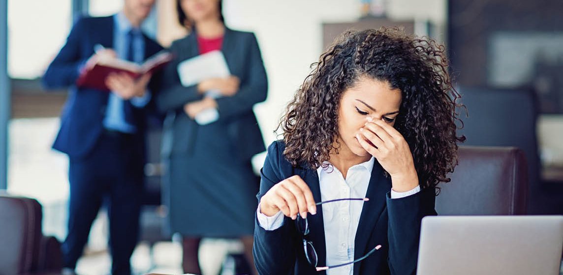 Stressed woman at desk with colleagues looking at her from a distance