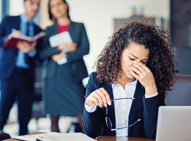 Stressed woman at desk with colleagues looking at her from a distance