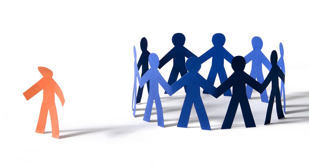 Orange paper person with dejected stance off to the side of a circle of blue paper people.