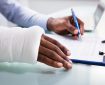 Overhead View Of Injured Man With Bandage Hand Filling Insurance Claim Form On Clipboard