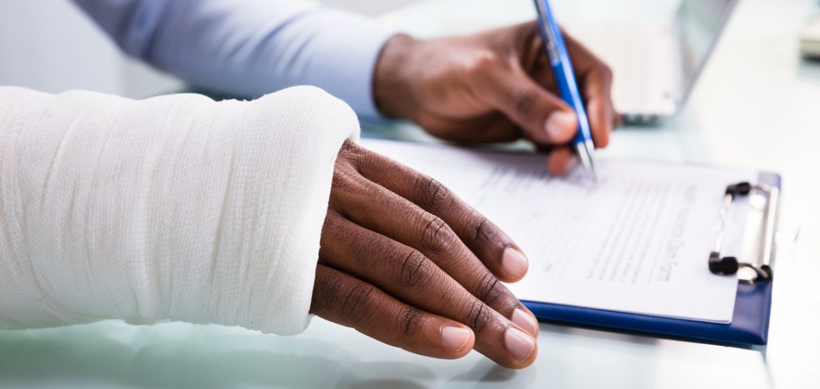 Overhead View Of Injured Man With Bandage Hand Filling Insurance Claim Form On Clipboard