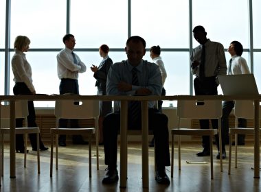 Businessman sits alone at his desk in the shadows while his colleagues stand in the background