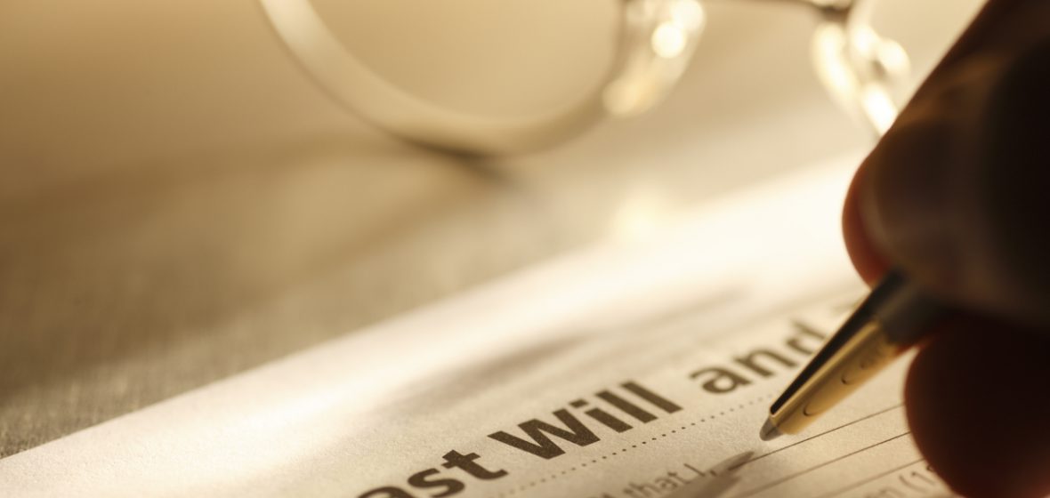 A close up of a man signing his last will and testament. A pair of eyeglasses rest on the desktop in the background.