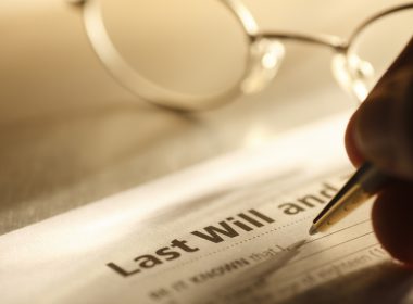 A close up of a man signing his last will and testament. A pair of eyeglasses rest on the desktop in the background.