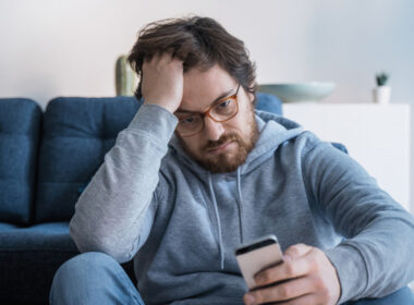 photo of man on couch looking sad and looking at mobile phone to show loneliness