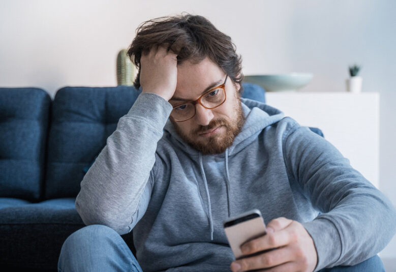 photo of man on couch looking sad and looking at mobile phone to show loneliness