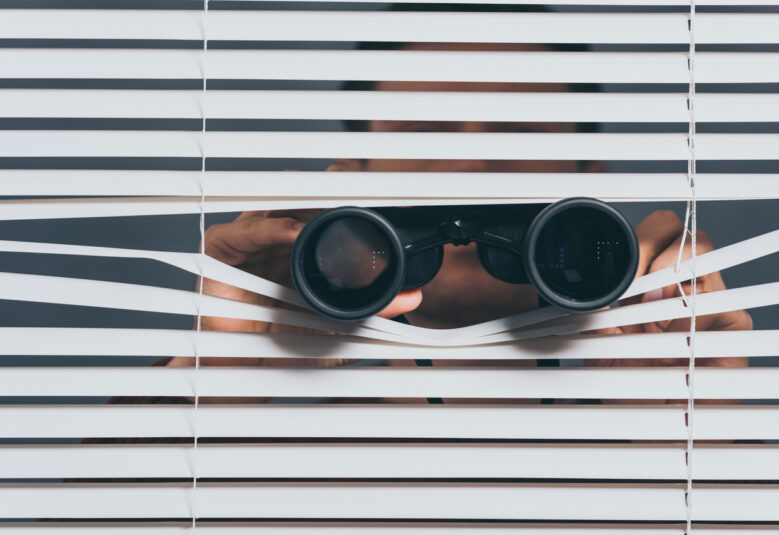 photo of person peering through blinds with binoculars to show surveillance